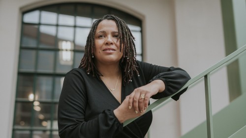 Tupoka lehnt an einem Treppengeländer. Sie hat ein schwarzes Oberteil an und steht vor einem Fenster.