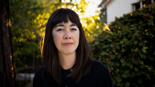 Jenny Odell trägt ein schwarzes Shirt und dunkle Haare. Im Hintergrund sind Büsche, Bäume und ein Haus zu erkennen.