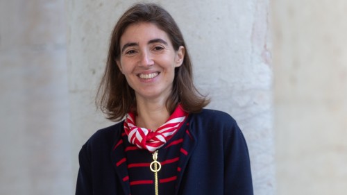 Chiara trägt schulterlange Haare, ein Halstuch und lächelt in die Kamera. Sie steht vor einer hellen Betonwand.