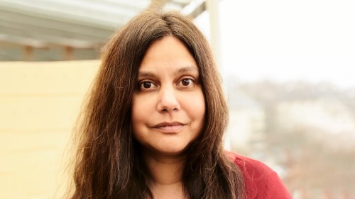 Mithu trägt lange dunkle Haare und ein rotes Tuch um den Oberkörper.