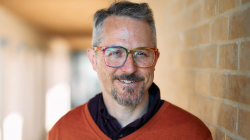 Sam Gregory trägt eine Brille, kurze Haare und Bart. Er trägt einen Pullover und darunter ein Hemd. Er lächelt in die Kamera und lehnt an einer Backsteinwand.