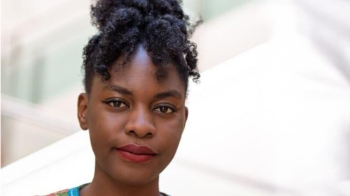 Foto von Esther vor hellem Hintergrund. Sie trägt ihre lockigen Haare hoch zusammengebunden und steht vor einem hellen Hintergrund.