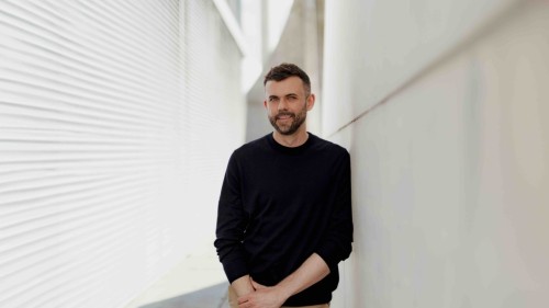 Ein Porträt von Janosch Delcker. Er trägt ein dunkles Sweatshirt trägt und lächelt in die Kamera, aufgenommen draußen in Berlin