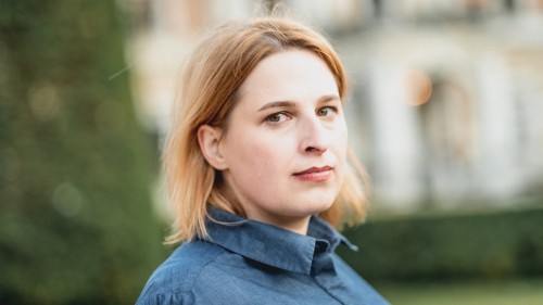 Natascha Strobl schaut seitlich in die Kamera. Im Hintergrund ist ein Gebäude und Baum sowie eine Wiese zu sehen. Natascha Strobl trägt eine Bluse und schulterlanges Haar