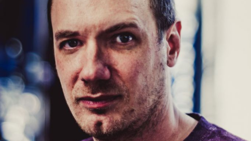 Portraitfoto von Christian Schiffer. Er trägt ein T-Shirt und kurze Haare. Er schaut skeptisch in die Kamera. Das Foto ist mit starkem Kontrast earbeitet. 