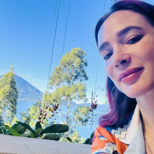 Woman in her 30s with pink hair and an orange shirt in front of mountains and a lake  