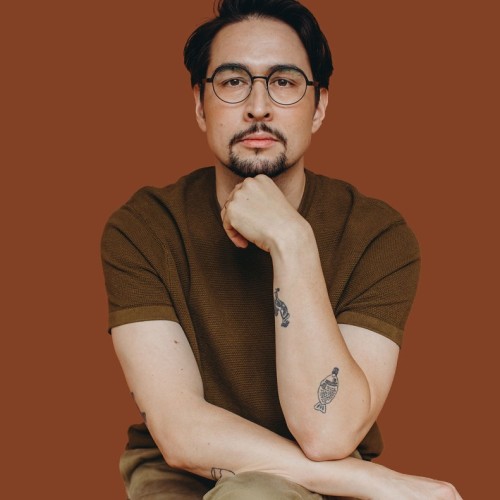 Eine Person sitzt vor einem einfarbigen, warm braunen Hintergrund. Sie trägt ein kurzärmliges, braunes Oberteil und eine beige Hose. Die Person hat mittellanges, dunkelbraunes Haar, eine runde Brille und einen kurzen Bart. Sie stützt den Kopf leicht auf eine Hand und blickt ruhig und direkt in die Kamera. Auf dem Unterarm sind zwei schwarze Tätowierungen sichtbar: ein Fisch und ein weiteres kleines Motiv. Die Haltung wirkt nachdenklich und entspannt.