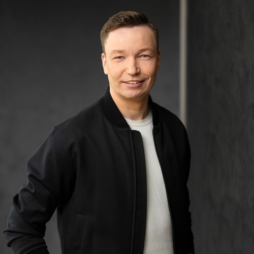 Porträt von Matthias Hölscher. Er lächelt und trägt eine schwarze Jacke über einem weißen T-Shirt