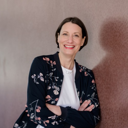Profilfoto von Jennifer Jones - sie trägt ein weißes Shirt, ein dunkles Blouson und lehnt mit verschränkten Armen vor der Wand. 