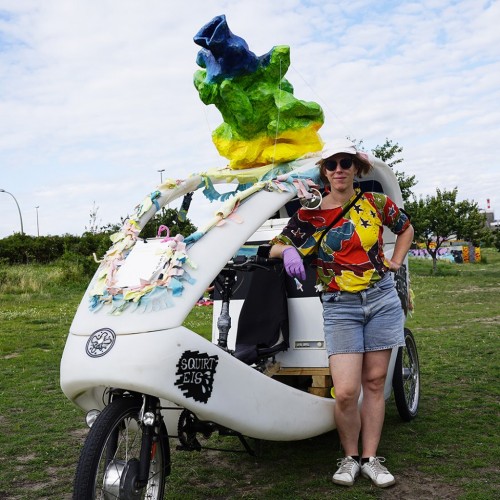 The performer Julia Frankenberg standing in front of a rikshaw with a huge prostate of a body with a female prostate remodelled after drawings from 1948.