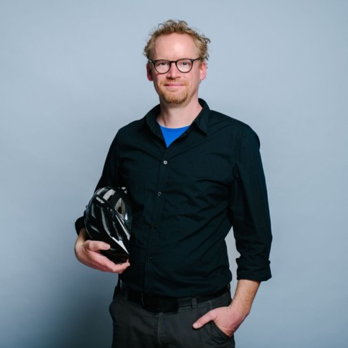 Portraitfoto Henning Krause: Ein weißer Mann Mitte vierzig mit Dreitagebart und Brille im schwarzen Oberhemd mit leicht hochgekrempelten Ärmel steht in der Mitte des Bilds und lächelt leicht in die Kamera. Unter dem rechten Arm hält er einen schwarzen Fahrradhelm.