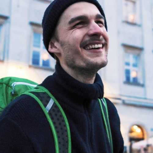 Foto von Jascha, Producer von Interscetion. Er lacht, hat einen drei Tage Bart und trägt einen dunkelblauen Wollpullover sowie eine dazu passende Mütze. Auf seiner Schulter ist ein grüner Rucksack. Er steht vor einer hellen Häuserwand.