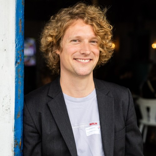 Ein Portraitfoto von Niklas Kossow, einem Mann mitte 30 mit blonden Locken, er trägt ein Lila TShirt und ein Sakko.