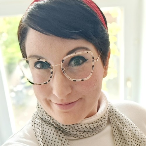 Eine Frau mit kurzen dunklen Haaren, einer getupften Brille und einem getupften Schal, sowie einem roten Haarreif vor einem Fenster.
