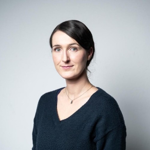 Portrait einer jungen blassen weißen Frau, sie trägt ihre dunklen Haare in einem Seitenscheitel und im Nacken zusammen gebunden. Sie trägt einen dunkelblauen Pullover mit V-Ausschnitt und eine silberne Kette mit einem Anhänger in Form einer Rakete. 