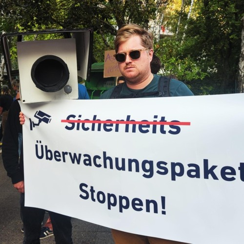 Foto: Demonstrationsszene gegen Überwachung. Ein Mann mit Sonnenbrille hält Plakat: 'Sicherheits' durchgestrichen, 'Überwachungspaket Stoppen!' nicht durchgestrichen. Daneben Person mit großer Pappkamera über dem Kopf.