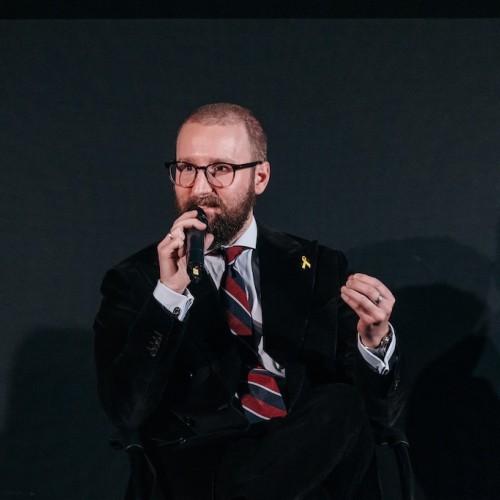 Auf dem Bild sitzt Monty Ott, hält ein Mikrophon in der einen Hand und erhebt die andere, um zu gestikulieren. Dabei drückt er Finger und Daumen zusammen. Der Blick ist aus der Betrachtenden-Perspektive nach links gerichtet.