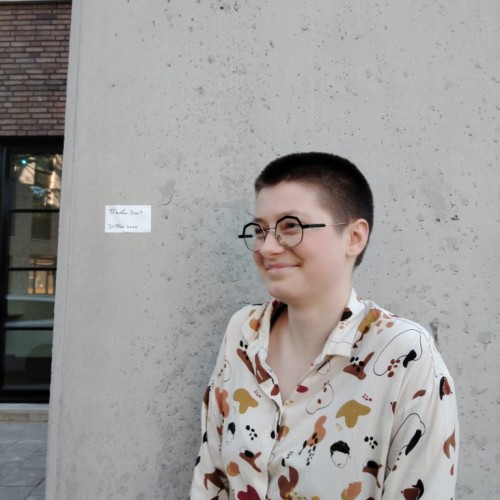 Junge Frau mit schwarzer Brille und braunem Buzzcut vor einem grauen Hintergrund. Sie trägt eine beige Blouse mit braun-schwarz geflecktem Muster.