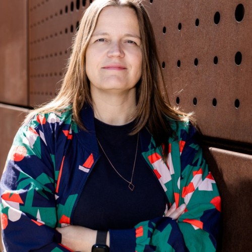 Eine Frau mit mittellangen Haaren lehnt an einer rost-roten Wand im Industrialdesign und schaut direkt in die Kamera. Sie hat die Arme vor der Brist verschränkt und trägt eine farbig gemusterte Jacke
