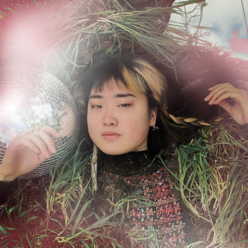 Alice holds a knowing expression as she lies on a bed of soil, surrounded by grass, reflective surfaces, and mystical red-white air