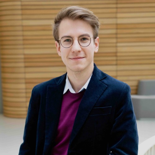 Das Bild zeigt einen jungen Mann mit Brille, der einen violetten Pullover sowie ein blaues Sakko trägt. 