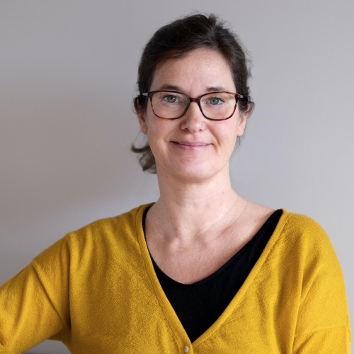Portrait-Foto von Anke Schöttler. Anke hat braune Haare, die sie zu einem Pferdeschwanz zusammen gebunden hat. Sie trägt eine Brille und einen gelben Pullover.