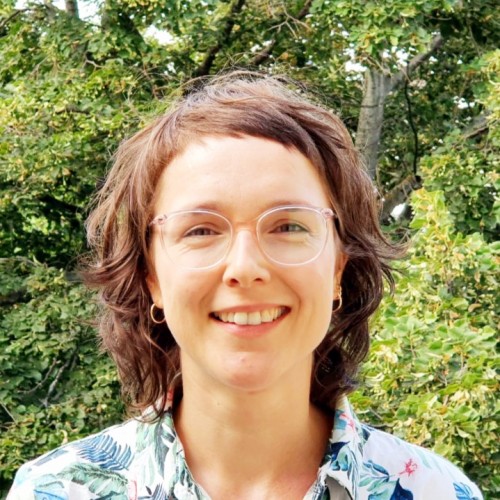 Porträt von Anna Spenn. Sie schaut lächelnd in die Kamera. Sie trägt eine rosafarbene Brille, ein weißes Oberteil mit Palmenblättern bedruckt und ihre braunen Haare offen. 