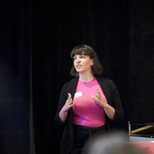 A white woman with brown hair, who is wearing a pink shirt and a black jacket, is speaking to an audience. She looks to one side. Her hands are in action, as she speaks. 