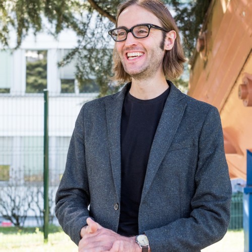 Physicist Dr. James Beacham wears a jacket and black glasses. 