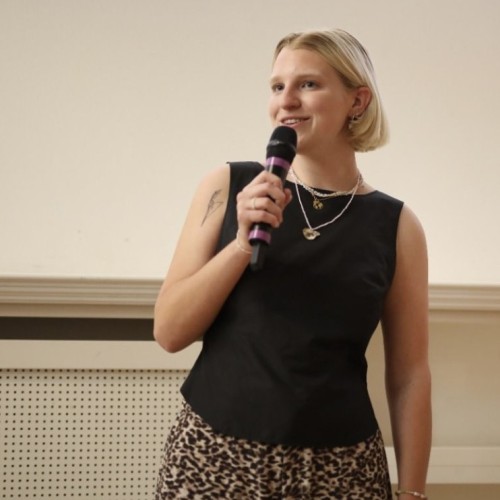 Eine Person spricht lächelnd in ein Mikro. Sie trägt ein schwarzes Oberteil und hat kinnlange, blonde Haare.