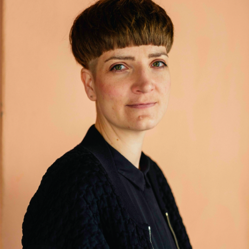 Sara Schurmann lächelt in die Kamera. Sie steht vor einer Apricot-farbenen Wand, trägt eine schwarze Blouson-Jacke über einer schwarzen Bluse und einen Pilz-Haarschnitt..