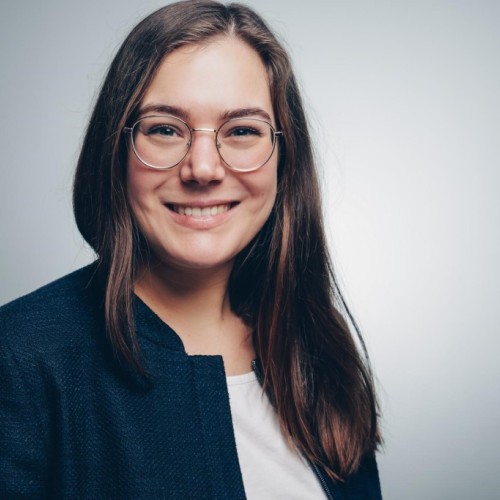 Frau mit langen braunen Haaren und goldener Brille vor grauem Hintergrund