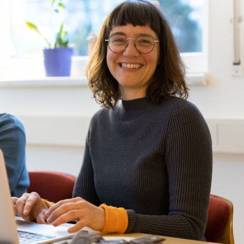 Simone am Schreibtisch vor einem Laptop. Sie lacht in die Kamera