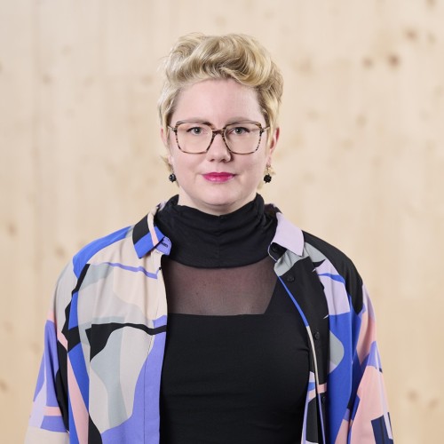 Frau mit blonden kurzen Haaren, Brille, Lippenstift, schwarzem Rollkragenpullover und buntem Hemd