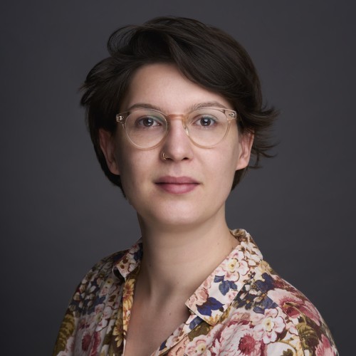 Portrait von Charlotte, sie steht in geblümter Bluse vor einer grauen Wand und trägt eine Brille