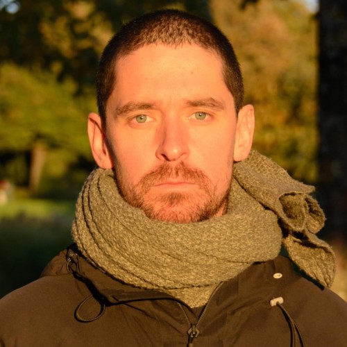 Image of a man with short black hair. He is wearing a scarf and a black jacket 