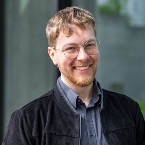 Ein Bild von Thomas Laschyk,  lächelnd vor einer Glaswand. Er trägt eine runde Brille, ein dunkelgraues Hemd und eine schwarze, samtartige Jacke. Im Hintergrund ist ein unscharfer, moderner Außenbereich mit Glas und etwas Grün zu erkennen