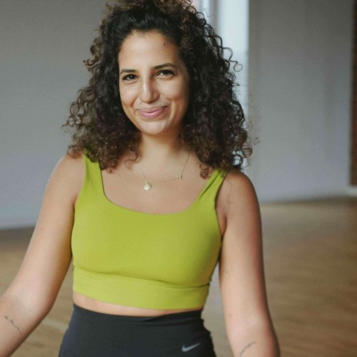 Dieses Bild zeigt eine Frau mit lockigem, dunklem Haar, das oben zu einem halben Dutt zusammengebunden ist. Sie trägt ein limettengrünes Sport-Top und eine schwarze, hochgeschnittene Sportleggings mit Nike-Logo. Auf beiden Armen sind feine Tätowierungen zu sehen. Sie sitzt im Schneidersitz auf einer hellen Matte in einem hellen Raum mit Holzboden. Im Hintergrund sind große Fenster und ein grauer bis weißer Wandbereich sichtbar. Die Frau lächelt entspannt in die Kamera und vermittelt eine freundliche, ruhige