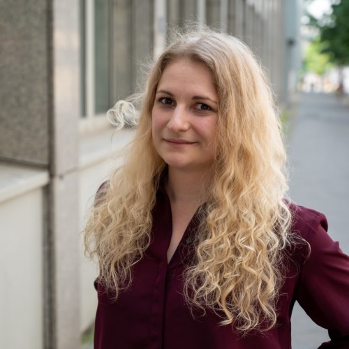 Eine Frau mit langen, lockigen, blonden Haaren und einem burgunderfarbenen Hemd steht draußen vor einem Gebäude. Sie schaut lächelnd in die Kamera. Im Hintergrund ist eine verschwommene städtische Straße mit Bäumen zu sehen.