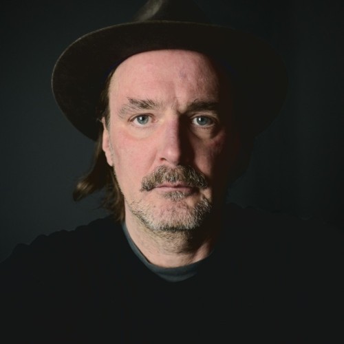 Ein (Selbst-) Portrait von Winson mit Hut vor dunklem Hintergrund. Das Licht kommt leicht seitlich rein, die rechte Gesichtshälfte ist dunkler als die linke.