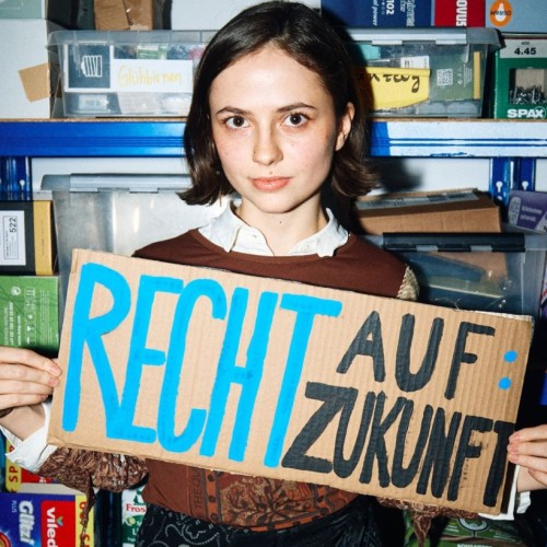Auf dem Bild ist eine junge Frau mit kurzen braunen Haaren zu sehen. Sie steht vor blauen Metallregalen, in denen verschiedene Kartons, Kisten und Haushaltsartikel lagern. Die Frau hält ein selbstgebasteltes Pappschild in den Händen. Auf dem Schild steht in großen, farbigen Buchstaben: „RECHT AUF: ZUKUNFT“. Die Frau schaut ernst, aber ruhig in die Kamera. Sie trägt eine weiße Bluse und darüber ein braunes,