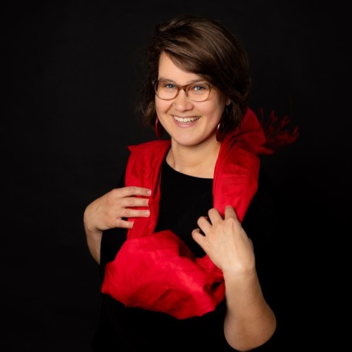 Dorothea blickt lächelnd in die Kamera. Sie hat kinnlange braune Haare und trägt eine beige Brille, rote Ohrringe, einen schwarzen Pulli und ein rotes Tuch.