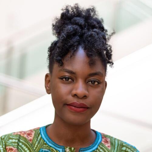 Esther Mwema, black African woman with curly afro hair in a bun, wearing green and red African material shirt, stands against blurred light background.