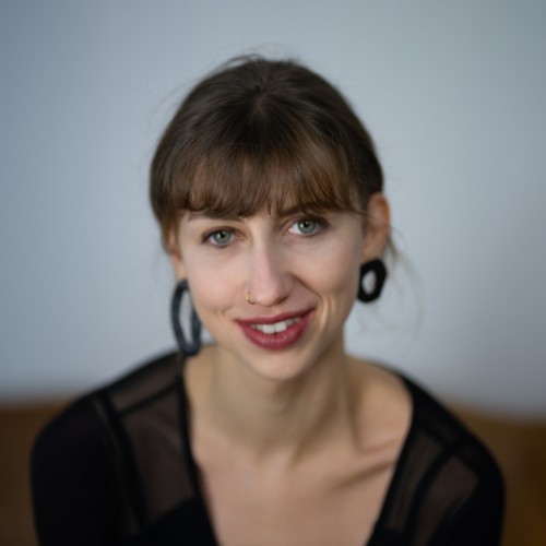 Porträtfoto von Fee. Sie hat einen Pony, zusammengebundene Haare, trägt ein schwarzes Oberteil aus Mesh und dazu schwarze große Ohrringe. 