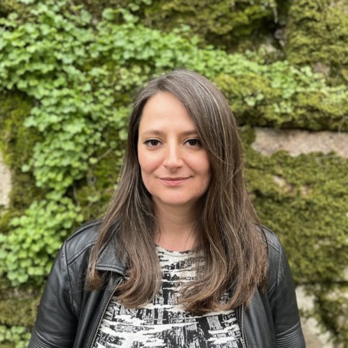 Foto einer Frau mit schulterlangen braunen Haaren, schwarzer Lederjacke, schwarz-weißem Kleid, vor einer bemoosten Felswand