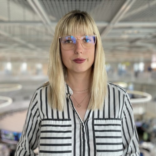 Eine junge Frau mit blonden Haaren, geradem Pony und schwarz-weiß gestreifter Bluse steht vor einem unscharfen Hintergrund. Sie trägt eine Brille und roten Lippenstift.