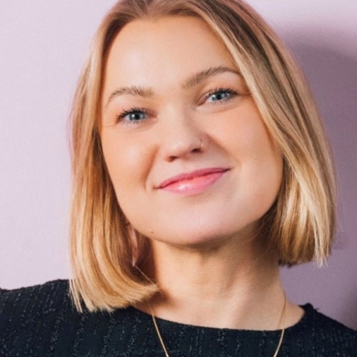 Eine junge Frau mit kinnlangem blondem Haar lächelt freundlich in die Kamera. Sie trägt ein schwarzes Oberteil mit feinem Muster und eine goldene Kette mit herzförmigem Anhänger. Der Hintergrund ist einfarbig in hellem Fliederton gehalten.