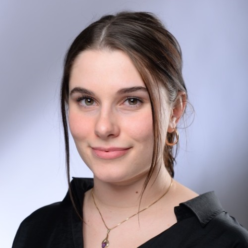 Eine junge Frau mit langen, dunkelbraunen Haaren, die teils offen und teils zurückgebunden sind, blickt leicht lächelnd in die Kamera. Sie trägt goldene Ohrringe, eine goldene Kette mit einem kleinen Anhänger sowie eine schwarze Bluse. Der Hintergrund ist neutral und hell.