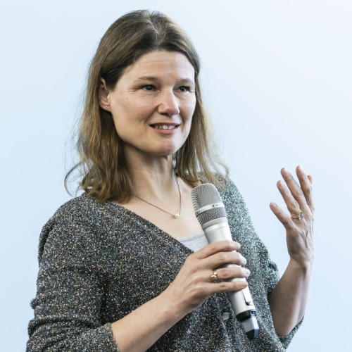 Eine Frau mit dunkelblonden Haaren hält ein Mikrofon in der Hand und spricht.