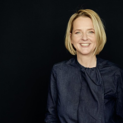 Schwarzer Hintergrund. Frau mit blondem halblangem Bob lächelt. Dunkelblaue Bluse.